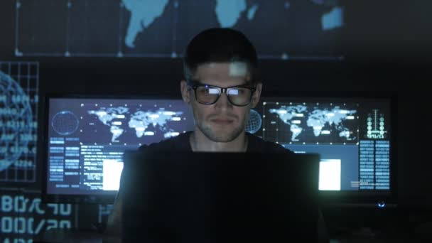 Хакер-програміст в окулярах працює на комп'ютері, а сині бінарні символи відображають його обличчя в центрі кібербезпеки, наповненому екранами . — стокове відео