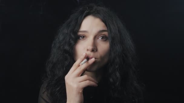 Портрет юной брюнетки с вьющимися волосами курит сигарету и смотрит в камеру на темном фоне — стоковое видео