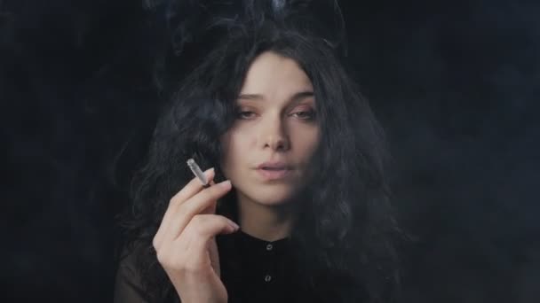 Portret van jonge brunette vrouw met krullend haar is roken van een sigaret en de camera te kijken op de donkere achtergrond — Stockvideo