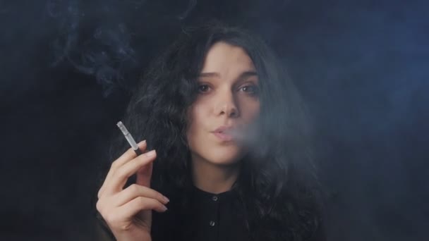Портрет юной брюнетки с вьющимися волосами курит сигарету и смотрит в камеру в темно-копченой комнате — стоковое видео