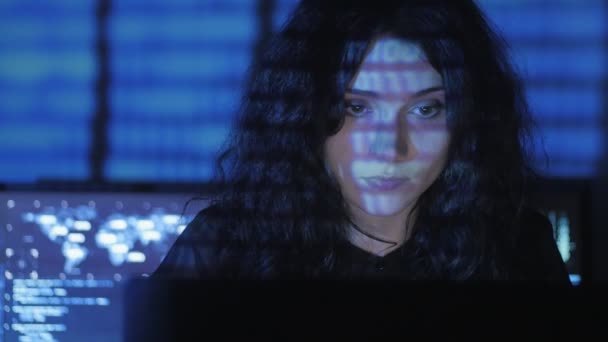 Woman Hacker programmer with curly hair is working on computer in cyber security center filled with display screens. Binary code on her face — Stock Video