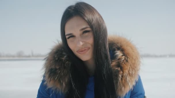 Portret van jonge vrolijke brunette vrouw blauwe jas glimlachen bij winter besneeuwde achtergrond — Stockvideo