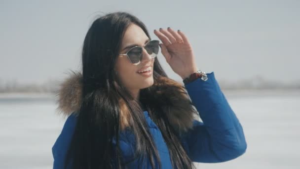 Portret van jonge mooie lachende brunette vrouw in zonnebril op winter sneeuwt achtergrond. — Stockvideo