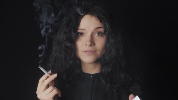 Portrait de jeune femme brune aux cheveux bouclés fume une cigarette et regarde la caméra sur le fond sombre — Video