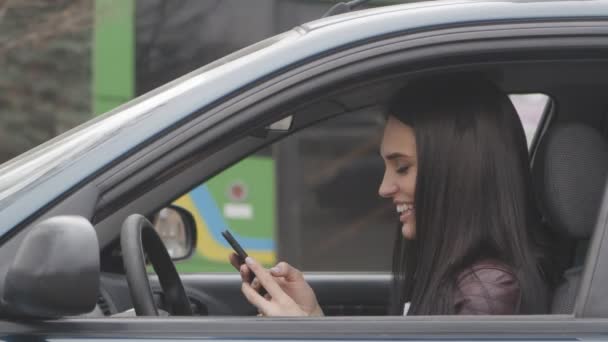 Joven hermosa mujer utiliza un teléfono inteligente mientras está sentado en un coche en la ciudad — Vídeos de Stock