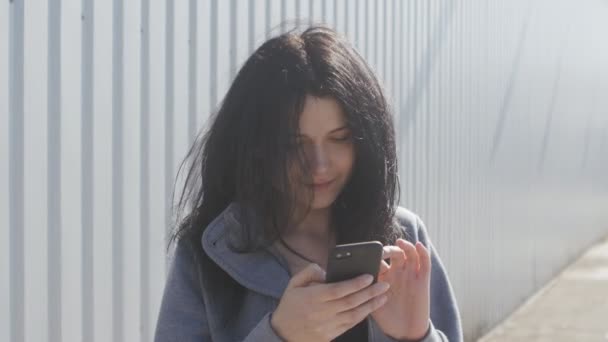 Porträt einer netten brünetten Frau, die ihr Smartphone draußen auf der Straße benutzt. der Wind weht leicht ihre Haare. — Stockvideo