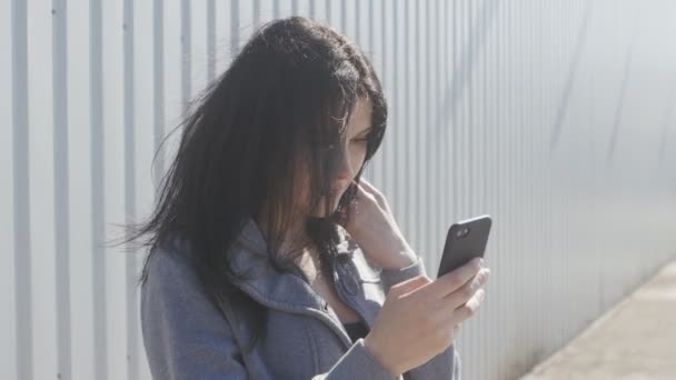 Portrait de femme brune mignonne utilisant smartphone en plein air dans la rue. Le vent agite facilement ses cheveux . — Video