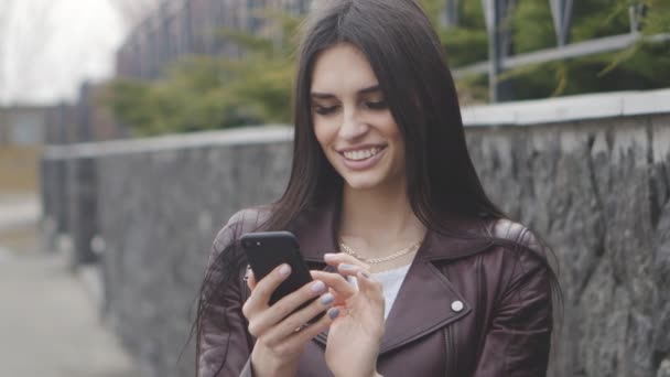 Porträt einer brünetten Frau, die auf der Straße lächelt und ihr Smartphone benutzt. — Stockvideo