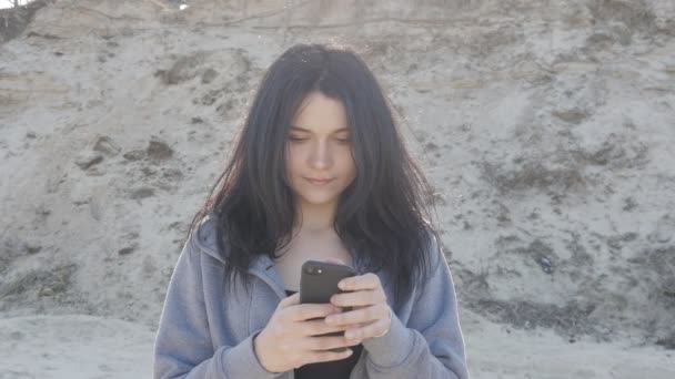 Портрет молодой женщины с использованием смартфона на открытом воздухе. Ветер легко машет ее волосами . — стоковое видео