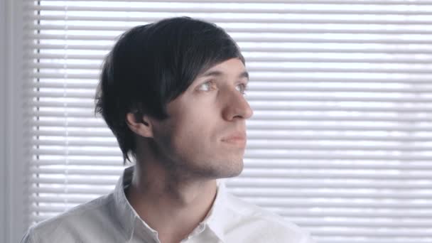 Portret van een jonge man in een wit overhemd op de achtergrond van de blinds. — Stockvideo