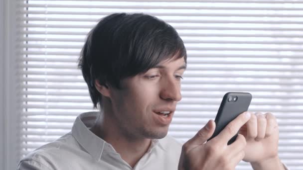 Retrato de un joven hombre de negocios con sonrisa utiliza un teléfono inteligente en el fondo de persianas — Vídeo de stock