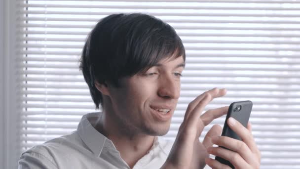Retrato de un joven hombre de negocios con sonrisa utiliza un teléfono inteligente en el fondo de persianas — Vídeo de stock