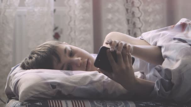 Jongen ligt in zijn slaapkamer in bed in de ochtend en maakt gebruik van een smartphone. — Stockvideo