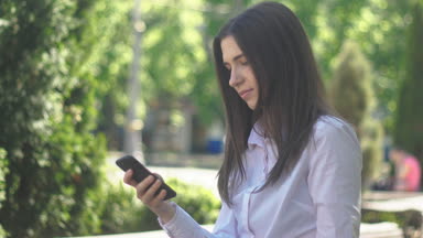Beyaz bir gömlek genç bir kadın portresi öğleden sonra şehirde bir sokakta bir akıllı telefon kullanır.