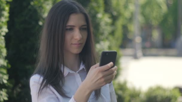 Retrato de una mujer joven con una camisa blanca utiliza un teléfono inteligente por la tarde en una calle de la ciudad . — Vídeo de stock