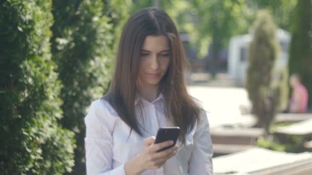 Portréja egy fiatal nő, egy fehér inget használ egy smartphone, délután egy utcában, a város. — Stock videók