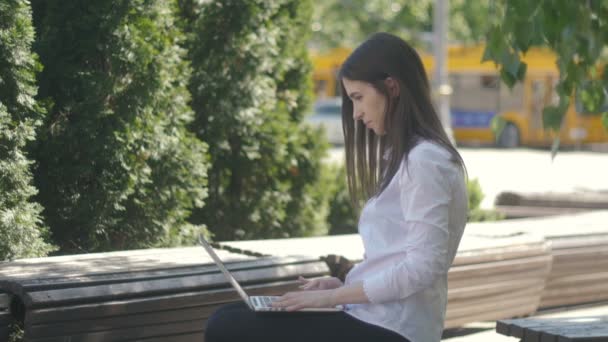 Młoda kobieta, wpisując na klawiaturze komputera laptopa siedząc na ławce na ulicy — Wideo stockowe