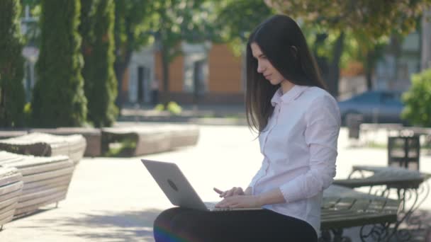 Ung kvinna skriver på bärbar dator tangentbord medan du sitter på bänken på gatan — Stockvideo