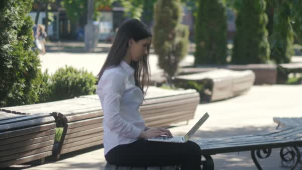 Młoda kobieta, wpisując na klawiaturze komputera laptopa siedząc na ławce na ulicy — Wideo stockowe