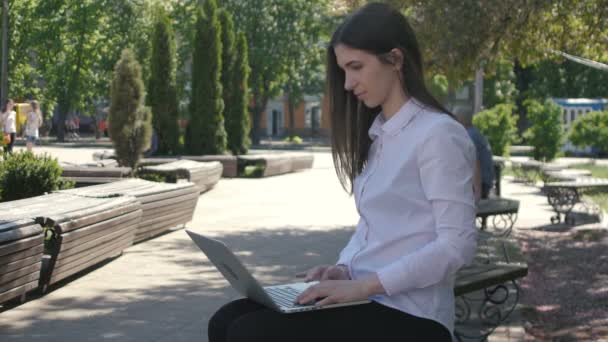 Młoda kobieta, wpisując na klawiaturze komputera laptopa siedząc na ławce na ulicy — Wideo stockowe