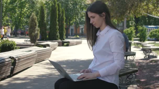 Junge Frau tippt auf Laptop-Computertastatur, während sie auf der Bank auf der Straße sitzt — Stockvideo