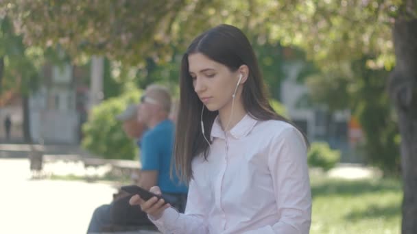 Vista lateral del retrato de mujer joven en camisa blanca escuchando música usando auriculares y teléfono inteligente. Al aire libre en ciudad — Vídeo de stock