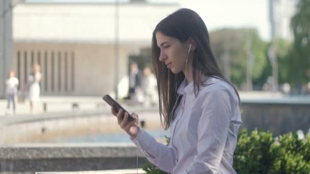 Vista lateral del retrato de mujer joven en camisa blanca escuchando música usando auriculares y teléfono inteligente. Al aire libre en ciudad — Vídeo de stock