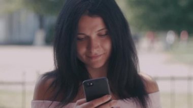 Onun akıllı telefon kısa mesaj okuma bir yaz parkta genç bayan portresi. Cep telefonu, açık kullanarak koyu saçlı güzel genç kız.