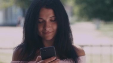 Onun akıllı telefon kısa mesaj okuma bir yaz parkta genç bayan portresi. Cep telefonu, açık kullanarak koyu saçlı güzel genç kız.