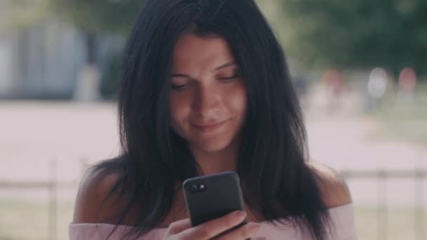 Portrait de jeune femme dans un parc d'été lisant un message texte sur son téléphone intelligent. Belle jeune fille aux cheveux foncés utilisant son téléphone portable, en plein air . — Video