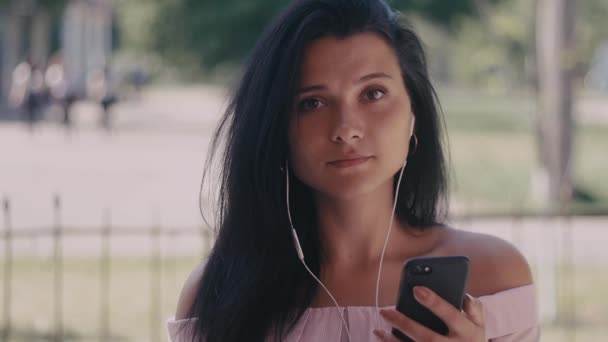 Feche a foto da jovem mulher ouvindo música no fone de ouvido enquanto usa o smartphone na cidade. Retrato tirado — Vídeo de Stock