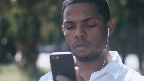 Retrato de hombre de negocios joven utiliza un teléfono inteligente y escucha música en los auriculares al aire libre en la ciudad — Vídeo de stock