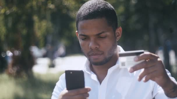 Şehir sokak arka plan üzerinde kredi kartı ile online alışveriş için akıllı telefon kullanan genç Afrika adam. Açık. — Stok video