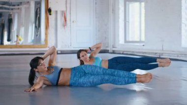 İki atletik kadın spor salonunda spor minderinin üzerinde İkili Bacak Kaldırma yapıyor. Spor kadınları egzersiz paspası üzerinde karın bölgesini çalıştırarak kaslarını geliştiriyor..