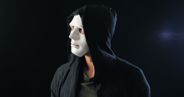 Anonymous man in a white mask covering his face and a black hood turns and looks at the camera. Black dark background. Concept of a member of a secret organization — Stock Video