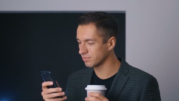 若い男性起業家はコーヒーを飲み、オフィスでスマートフォンを使用します。ハンサムな笑顔のビジネスマンはコーヒーブレイク中に彼の携帯電話を使用します。. — ストック動画