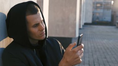 Mahalledeki genç gangster uyuşturucu satıcısı portresi açık şehir arka planında akıllı telefon kullanıyor.