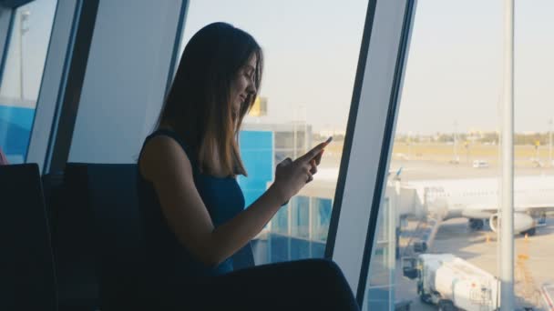 Viajera joven utiliza un teléfono inteligente en una terminal del aeropuerto — Vídeos de Stock