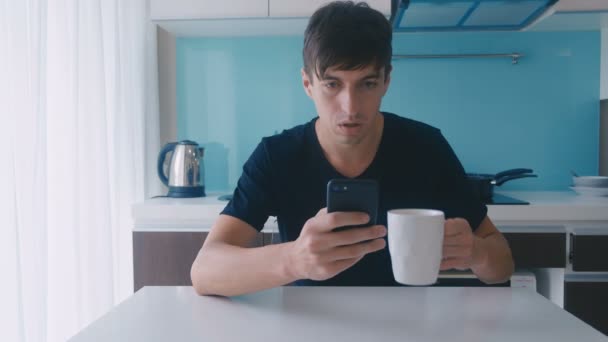 Hombre se sorprende por el mensaje en el teléfono inteligente y escupe café en la mesa en casa en la cocina — Vídeos de Stock