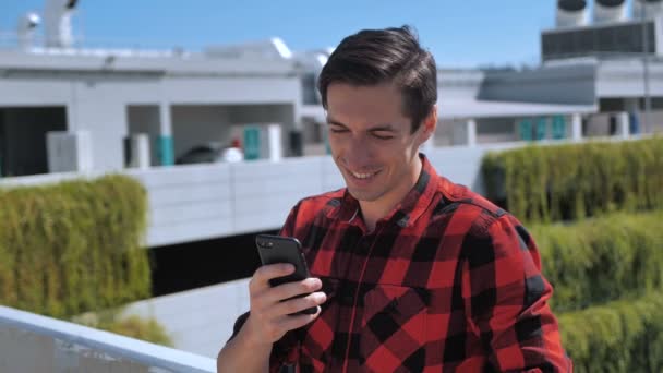 Joven hombre sonriente en camisa a cuadros mensaje de texto en el teléfono moderno teléfono inteligente en el techo en el fondo de la ciudad urbana — Vídeos de Stock