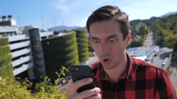 Hombre guapo en camisa a cuadros sosteniendo el uso de teléfono móvil inteligente sorprendido cara emocional mostrando el éxito mantener la mano puño de pie techo hacer sí victoria pulgares hacia arriba signo de la mano — Vídeos de Stock