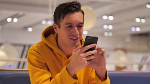 Junger lächelnder glücklicher Mann in gelbem Kapuzenpulli mit Smartphone in gemütlichem Café zur Abendzeit. — Stockvideo