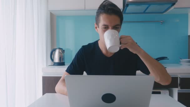 Joven hombre de negocios freelancer que trabaja en el ordenador portátil y beber café en la cocina casera . — Vídeo de stock