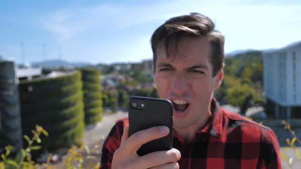男はスマートフォンで読んだメッセージに腹を立てている。都市の背景にスマートフォンの屋外を使用して再生シャツで積極的な若い男. — ストック動画