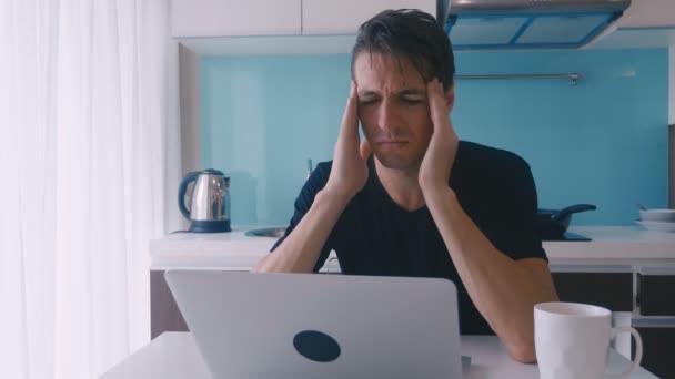 Joven deprimente sufre y tiene un dolor de cabeza mientras trabaja en un ordenador portátil en casa en la cocina . — Vídeo de stock