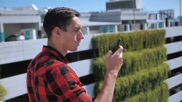 Atrakcyjny mężczyzna w koszuli w kratę za pomocą smartfona dotykającego ekranu na zewnątrz w tle miejskiego miasta. Urządzenia, ludzie i współczesna koncepcja młodzieży — Wideo stockowe