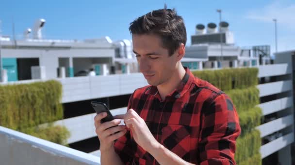 Atrakcyjny mężczyzna w koszuli w kratę za pomocą smartfona dotykającego ekranu na zewnątrz w tle miejskiego miasta. Urządzenia, ludzie i współczesna koncepcja młodzieży — Wideo stockowe