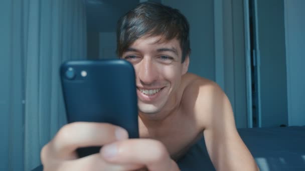 Bonito nu sorrindo homem deitado na cama em casa e usando smartphone — Vídeo de Stock