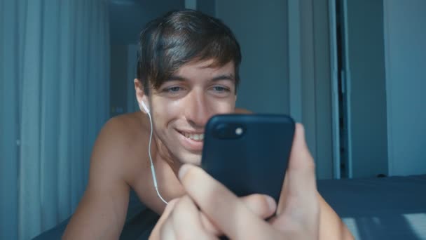 Bonito homem sorrindo nu com fones de ouvido deitado na cama em casa e rolando no smartphone — Vídeo de Stock