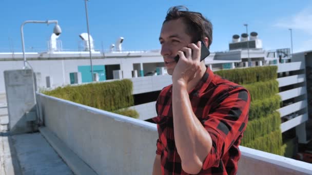 Uśmiechnięty młodzieniec w koszuli w kratę rozmawiający przez telefon komórkowy na dachu budynku na tle miasta — Wideo stockowe